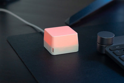 A small cube-shaped electronic device with a glowing pink top half and a white bottom half, placed on a dark desk next to a keyboard. A white cable extends from the left side.